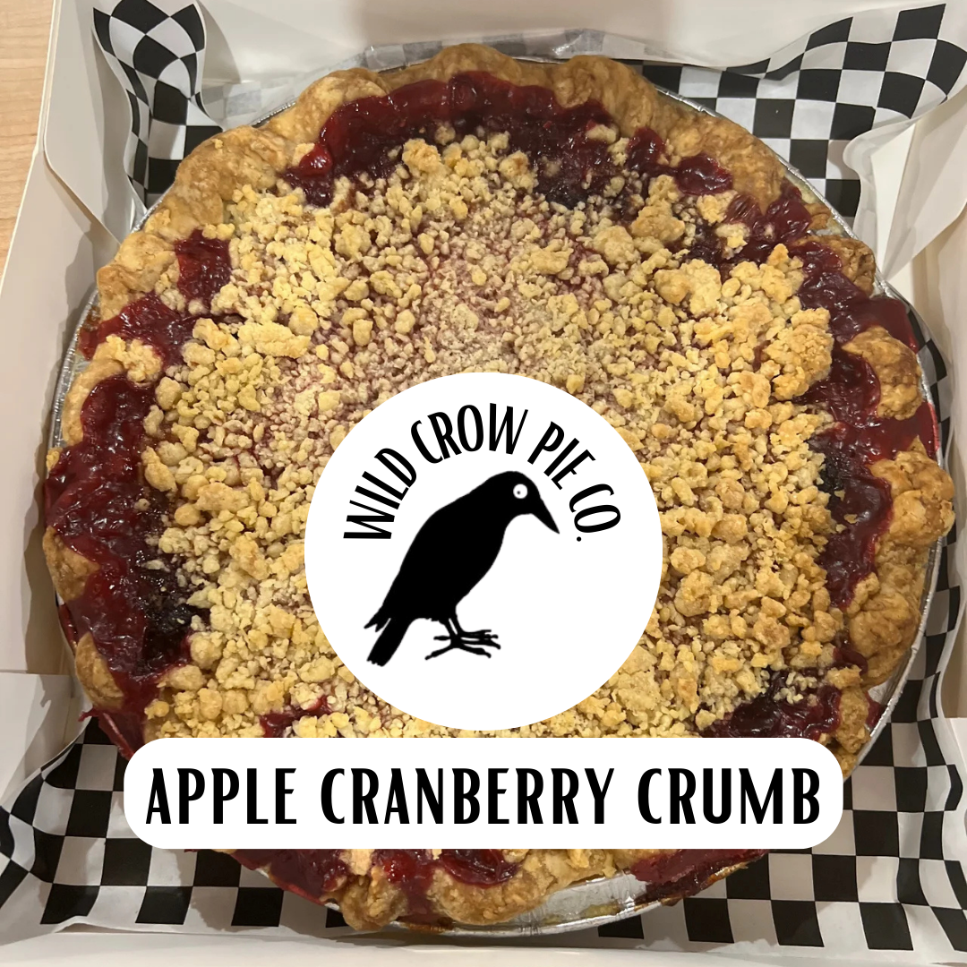 A baked apple cranberry crumb pie topped with crumbs and a piece of paper with 'Wild Crow Pie Co.' and 'APPLE CRANBERRY CRUMB' written on it, placed on a checkered patterned surface.