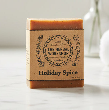 Bar of soap with 'The Herbal Workshop' branding on a marble surface with towels and a plant in the background.