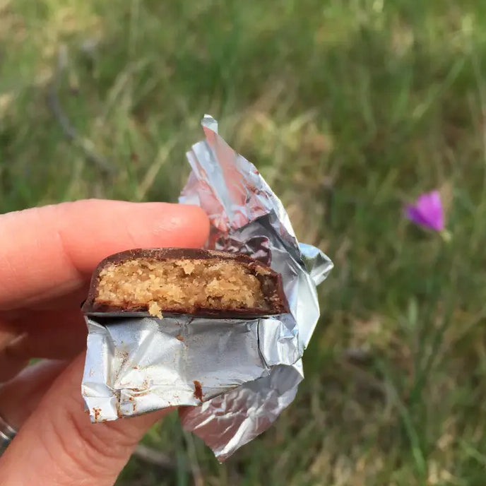 grass in background with hand holding a silver foil-wrapped half-eaten candy that is a crumbly tan covered in brown chocolate