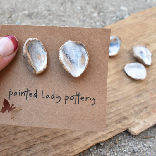 A set of ceramic earrings shaped like oyster shells, held in a person's hand with a small brown card labeled 'Painted Lady Pottery'.