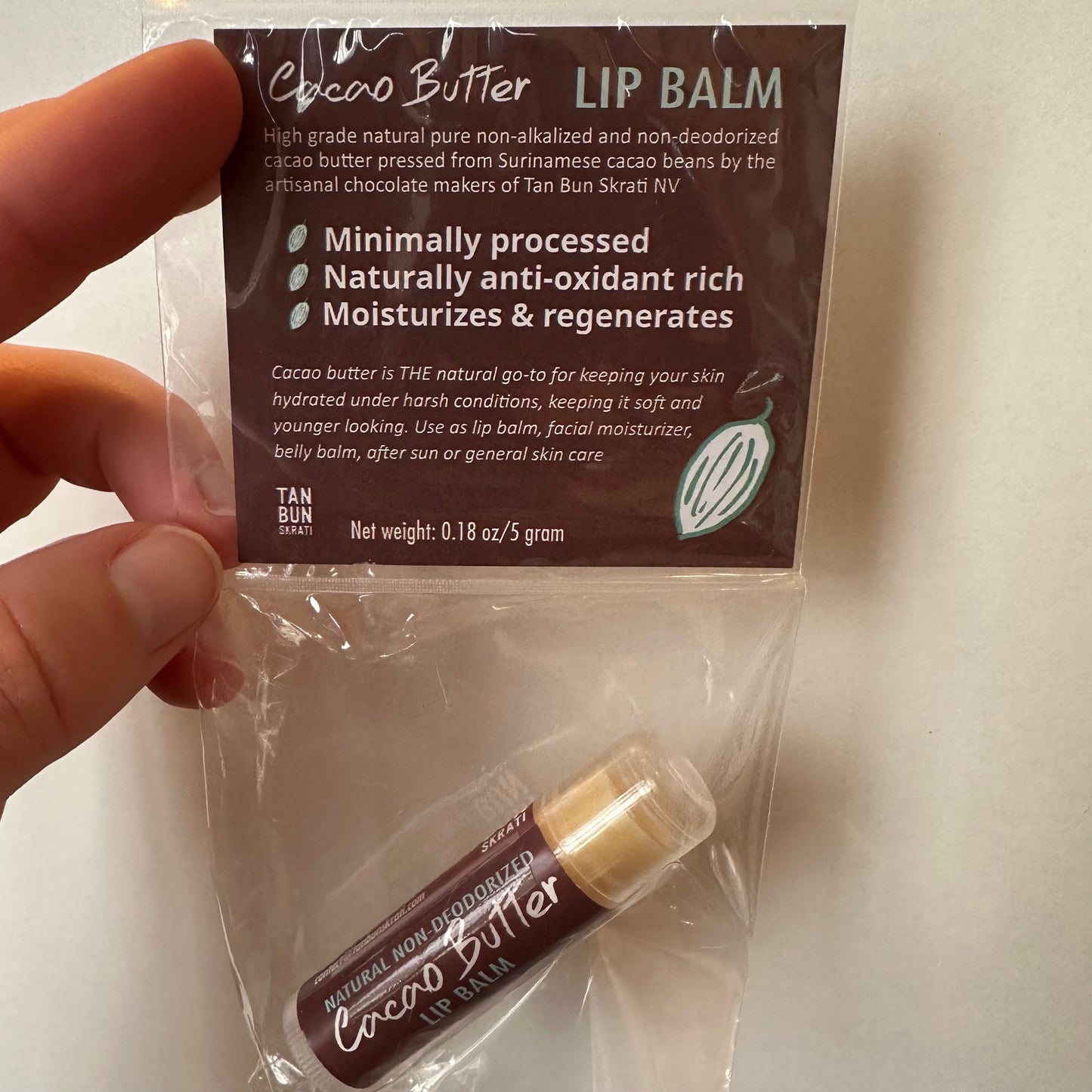 A hand holding up a small plastic bag containing tube of lip balm with brown label that says “Cacao Butter” - the bag has a brown label with a lot of information, some of which is “Cacao Butter LIP BALM” “Minimally processed” “Naturally anti-oxidant rich” “Moisturizes & regenerates”