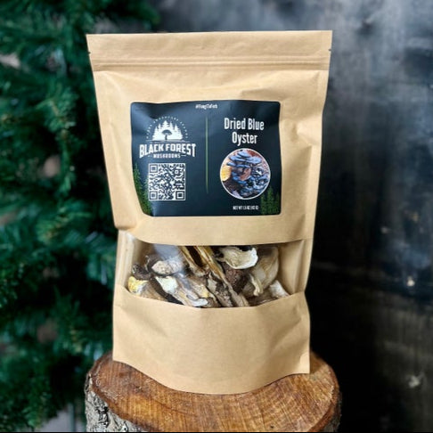 Brown bag of dried mushrooms with black label saying "BLACK FOREST" "Dried Blue Oyster" with an evergreen tree in the background