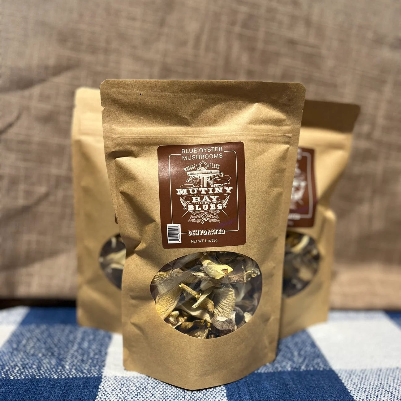 Kraft paper packaging with a clear window showing dried mushrooms on a blue and white checkered tablecloth.