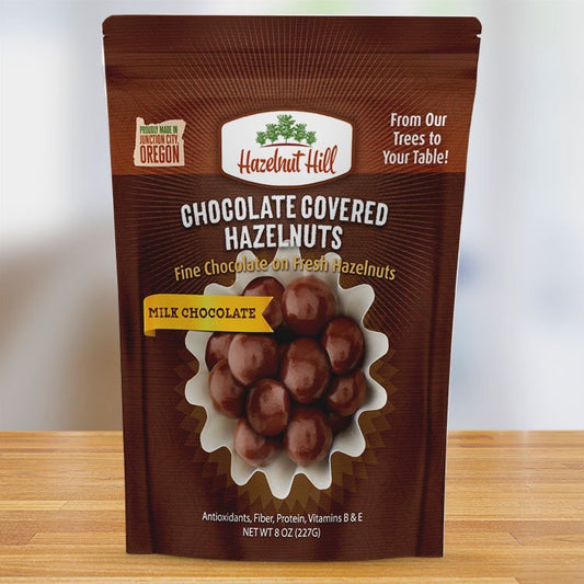A package of Hazelnut Hill milk chocolate covered hazelnuts. The packaging is brown with a transparent window showing the chocolate-covered hazelnuts inside.
