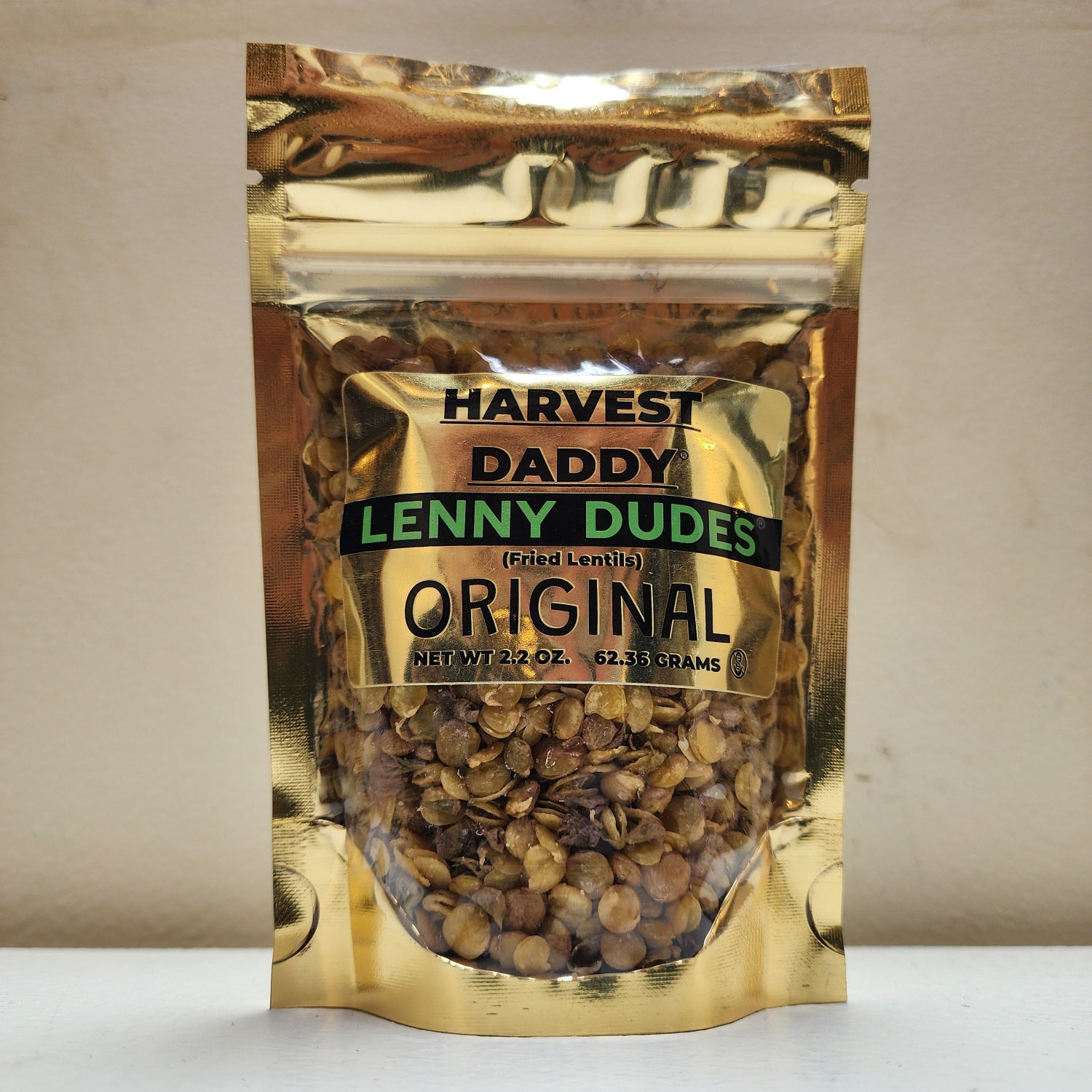 A bag of Harvest Daddy Lenny Dudes (fried lentils) in a shiny gold resealable pouch, shown on a light beige surface.