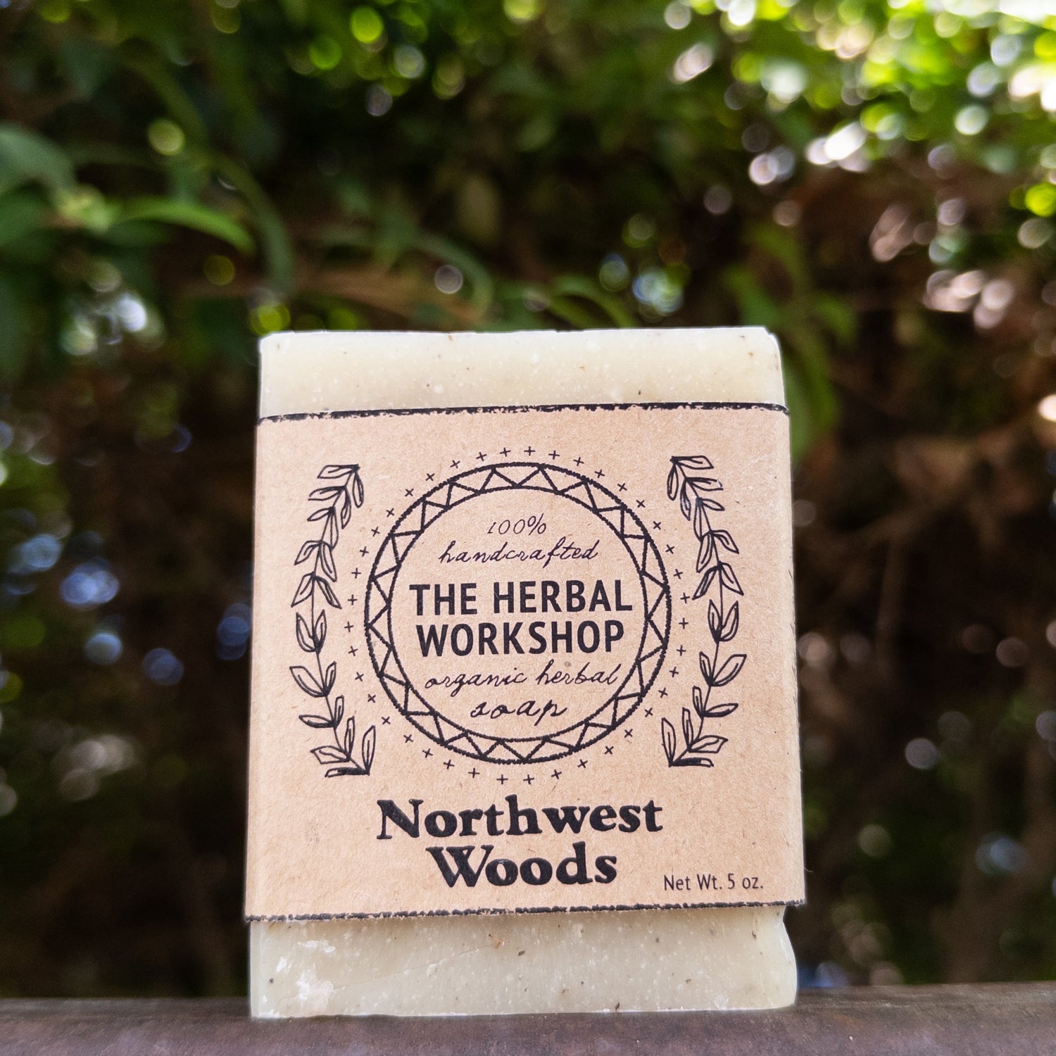 A rectangular bar of soap standing upright outdoors on a dark surface, wrapped in a kraft paper label that reads 'The Herbal Workshop' and 'Northwest Woods.' The background is a blurred green outdoor setting with sunlight and bokeh.