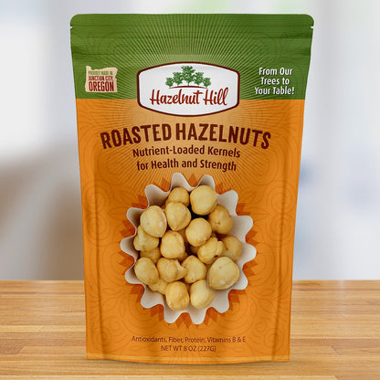 Packaging of Hazelnut Hill Roasted Hazelnuts showing the product through a clear window, with green and orange colors and text indicating the product is nutrient-loaded for health and strength, from our table! Also mentioning it's a product of Oregon.