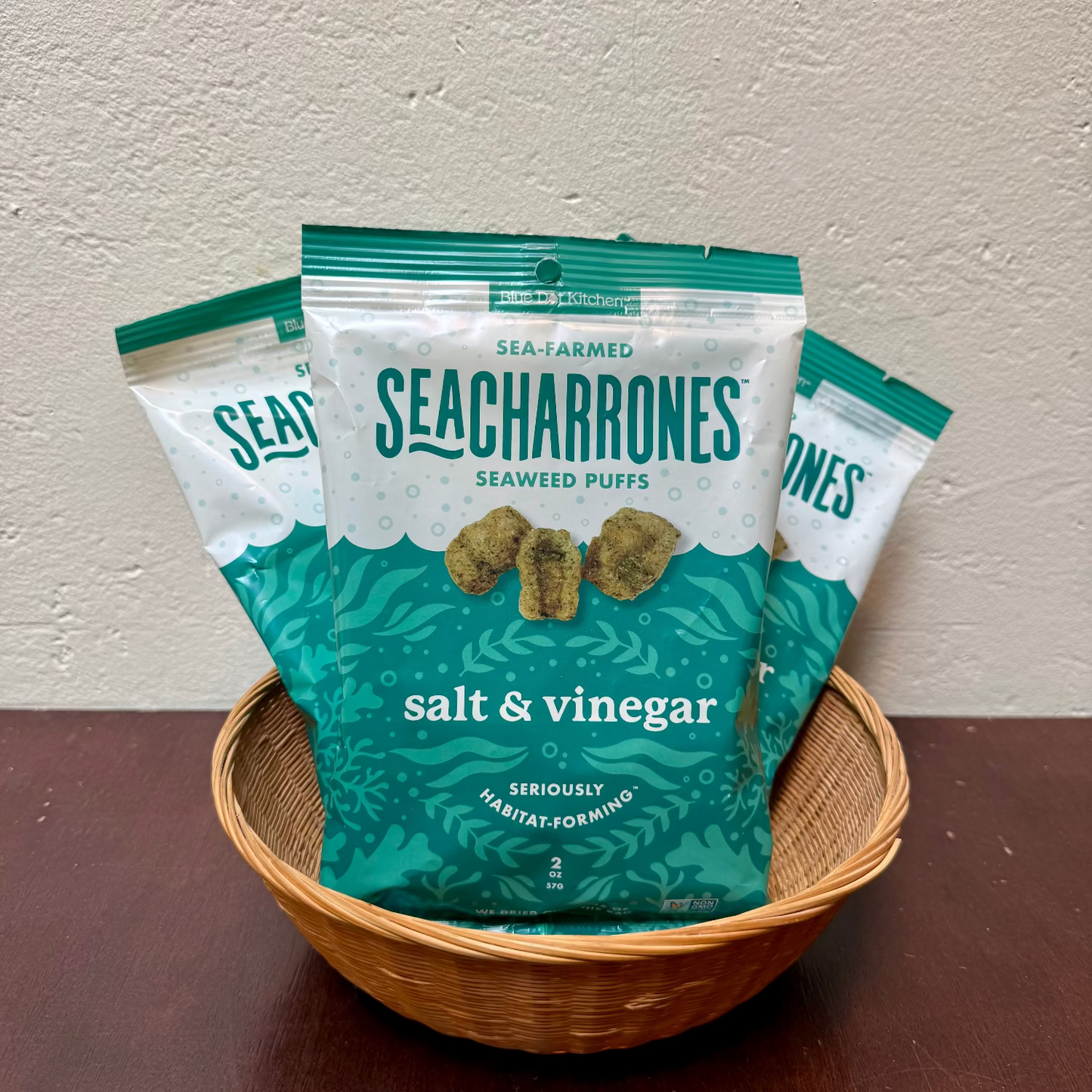 Three bags of Seacharrones seaweed puffs in the salt & vinegar flavor, arranged in a small woven basket on a dark wooden surface against a textured beige wall.