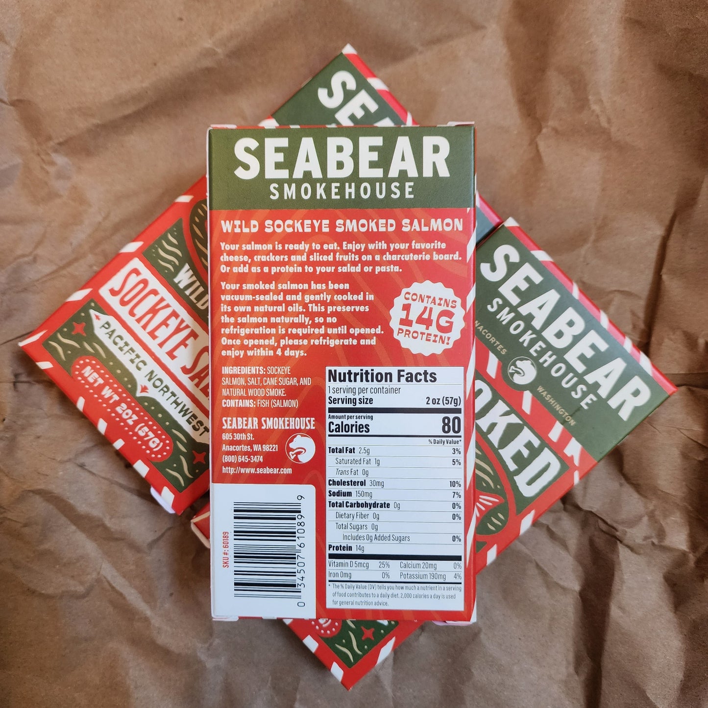 Three Seabear Smokehouse wild sockeye smoked salmon snack boxes arranged on a crumpled brown paper background. The center box is shown upright, displaying the green and white brand name, product type, and nutrition facts panel, while two additional boxes are partially visible underneath.