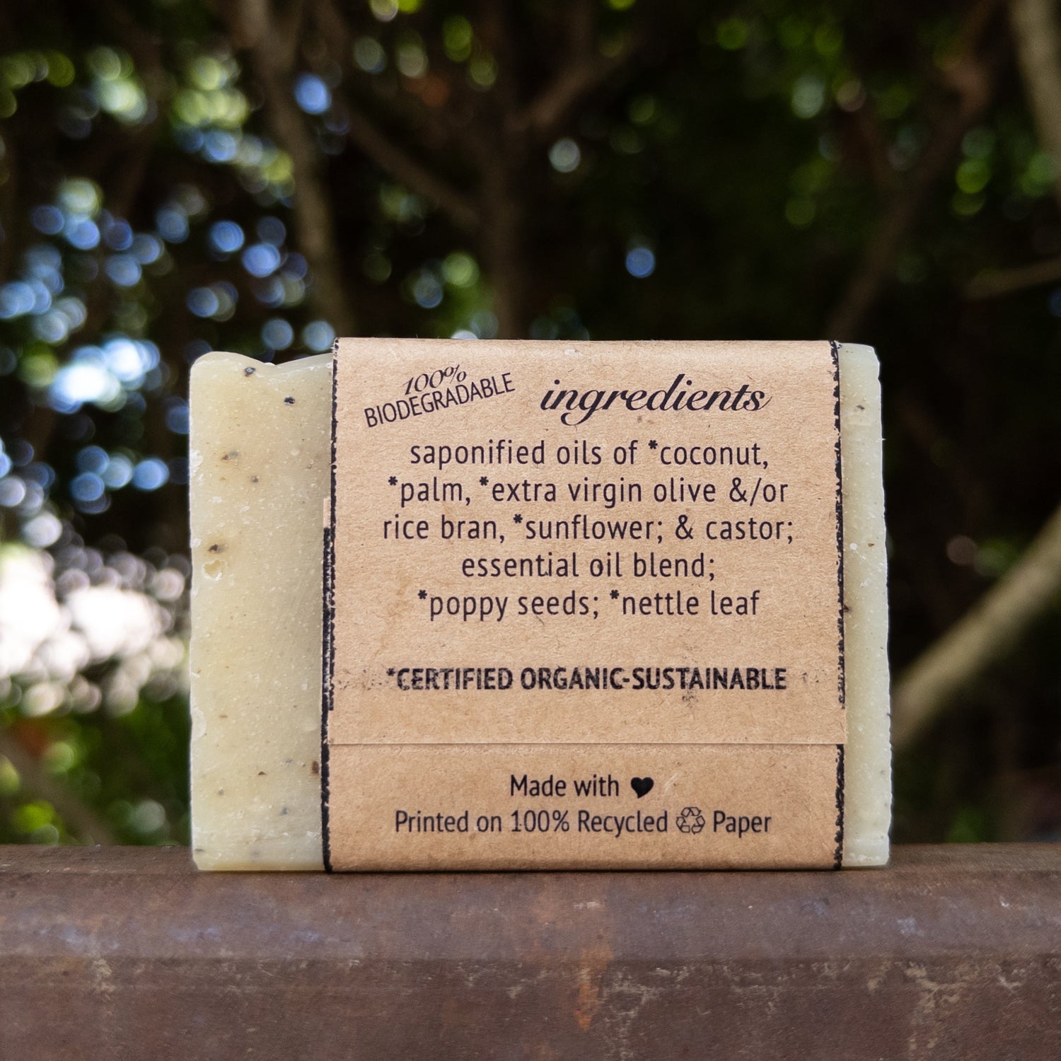 A rectangular bar of handmade soap standing upright on a wooden surface, with a paper band around the middle showing ingredient text. The soap is photographed outdoors with blurred green foliage in the background.