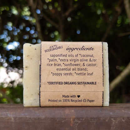 A rectangular bar of handmade soap standing upright on a wooden surface, with a paper band around the middle showing ingredient text. The soap is photographed outdoors with blurred green foliage in the background.