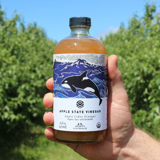 Glass bottle of orange-brown liquid held by a hand in front of a background of green leafy trees. Bottle has image of an orca whale in front of blue waves and says APPLE STATE VINEGAR Apple Cider Vinegar