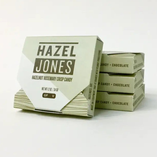 small light green thin square boxes, three stacked with one leaning up against them, text on the front: "HAZEL JONES / Hazelnut Rosemary Crisp Candy / GF V'