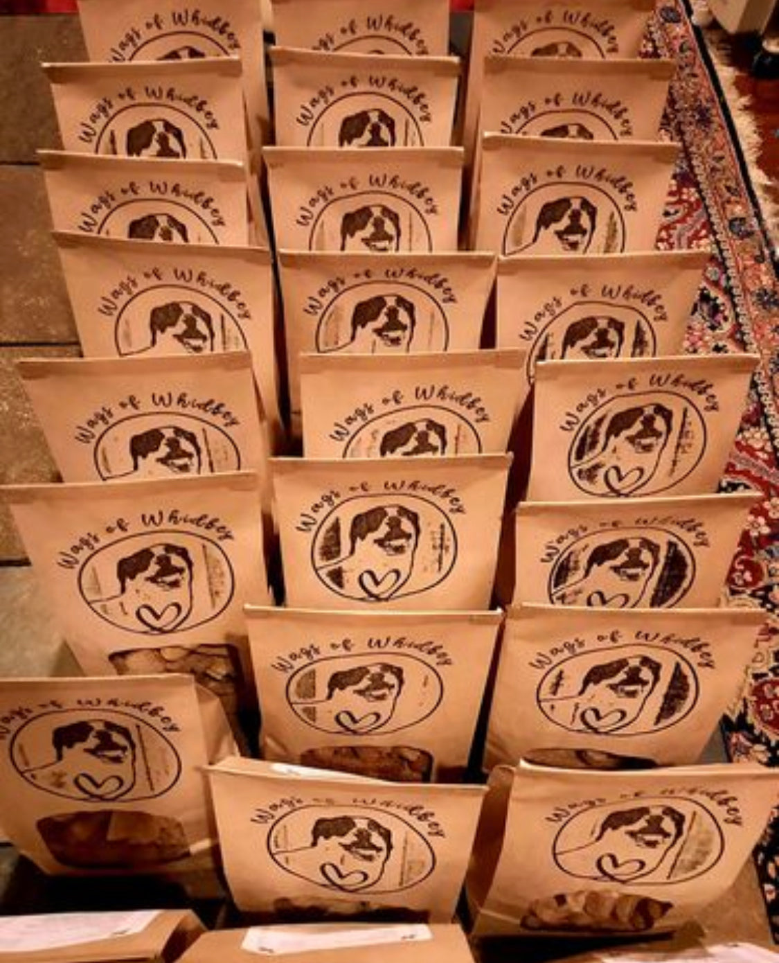 
                  
                    Rows of small paper bags filled with Wags of Whidbey dog treats, on a red paisley carpet
                  
                