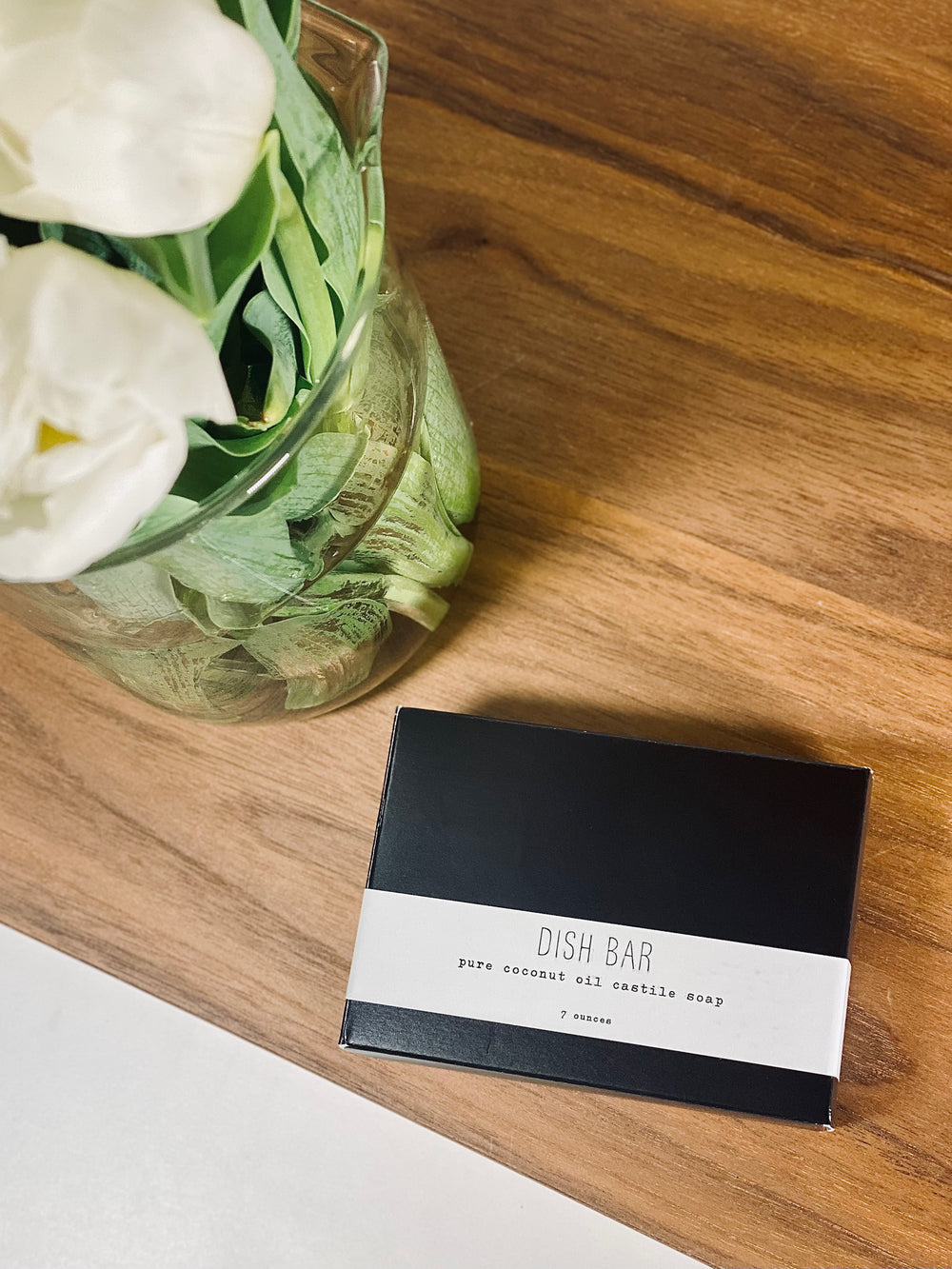 Black small box of bar soap on a wooden table next to a white flower in a vase